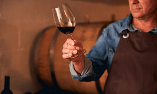 un produttore di vino tiene in mano un calice di rosso, dietro si intravede una botte di legno